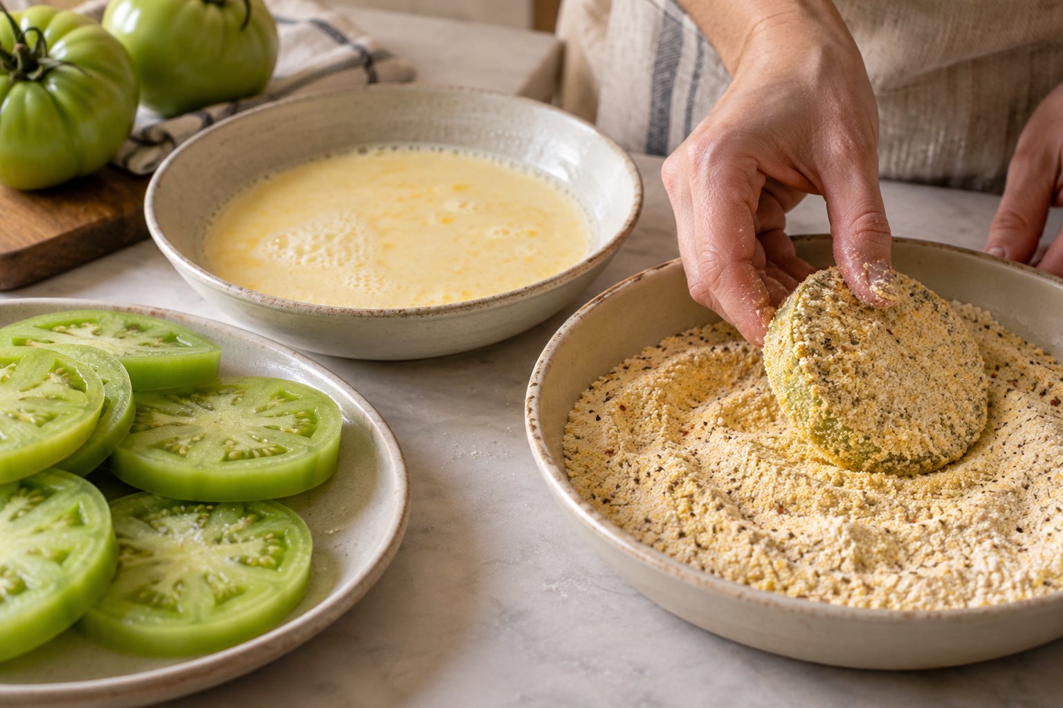 A tomato round pressed into cornmeal coating, with egg-buttermilk wash in a second shallow dish and uncoated green rounds beside it.